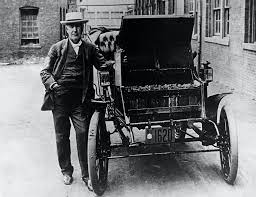 در یک عکس سیاه و سفید، مخترع توماس ادیسون در کنار یک موتور الکتریکی بسیار قدیمی ایستاده است.